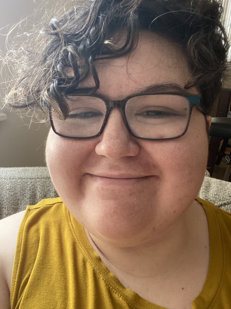 A close-up selfie of Margaret “Peggy” Wright, a smiling person with short, curly dark hair and glasses, wearing a mustard-yellow shirt. They appear warm and friendly, looking directly at the camera.